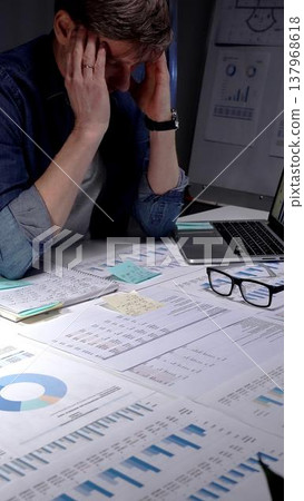 Young businessman with glasses sits at a dark office desk late at night, overwhelmed by documents and a tablet, clutching his head from stress, burnout and exhaustion Young businessman with glasses sits at a dark office desk late at night, overwhelmed by documents and a tablet, clutching his head from stress, burnout and exhaustion 137968618