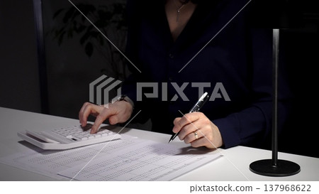 Woman calculating numbers on calculator, reviewing financial documents, working late in dimly lit office. Audit and taxes in business 137968622