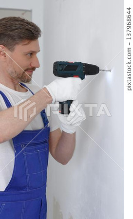 Professional male construction worker in a blue jumpsuit, safety goggles, and work gloves is screwing a screw into the wall using a cordless screwdriver, carrying out a careful home renovation 137968644
