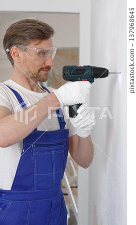 Professional male construction worker in a blue jumpsuit, safety goggles, and work gloves is screwing a screw into the wall using a cordless screwdriver, carrying out a careful home renovation Professional male construction worker in a blue jumpsuit, safety goggles, and work gloves is screwing a screw into the wall using a cordless screwdriver, carrying out a careful home renovation 137968645