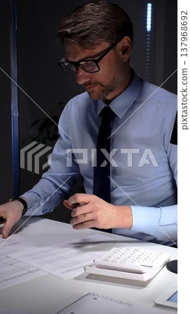 Businessman diligently auditing financial documents with a magnifying glass under focused lamp light. Audit and taxes in business Businessman diligently auditing financial documents with a magnifying glass under focused lamp light. Audit and taxes in business 137968692
