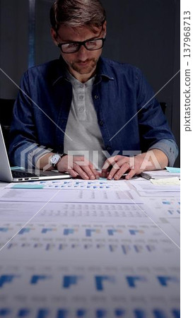 Professional man in glasses working late at night, analyzing financial data on a digital tablet, managing business reports, and extending productivity in a dark office environment Professional man in glasses working late at night, analyzing financial data on a digital tablet, managing business reports, and extending productivity in a dark office environment 137968713