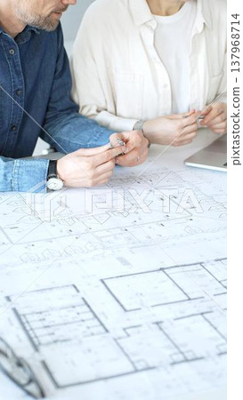 Engineering professionals collaborating on an architectural blueprint, using a compass to design a modern building, planning construction with a house model and laptop 137968714