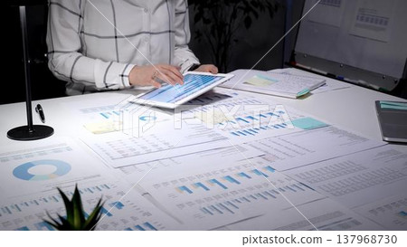 Business professional analyzing financial data and market trends on a digital tablet at a desk filled with various charts, graphs, and reports, working on corporate strategy 137968730