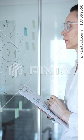 Professional woman in glasses working on business strategy, analyzing data on a clipboard, and sketching a mind map on a clear glass whiteboard in a modern office Professional woman in glasses working on business strategy, analyzing data on a clipboard, and sketching a mind map on a clear glass whiteboard in a modern office 137968950