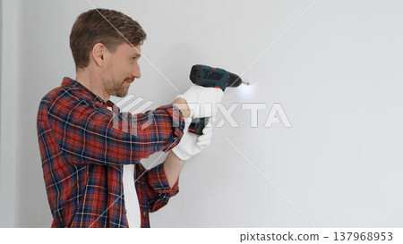 Middle aged male worker wearing red checked shirt and white gloves, is drilling hole in a wall with cordless drill during home renovation work. Portrait view Middle aged male worker wearing red checked shirt and white gloves, is drilling hole in a wall with cordless drill during home renovation work. Portrait view 137968953