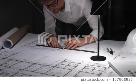 Architect working late, drawing blueprint details on a building floor plan design with a lamp illuminating the table. Business and architect people concept Architect working late, drawing blueprint details on a building floor plan design with a lamp illuminating the table. Business and architect people concept 137968955