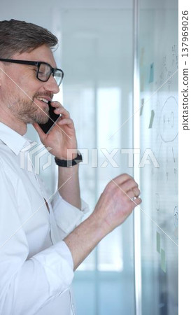 Professional businessman in glasses communicating on smartphone, standing by a modern glass whiteboard with notes, brainstorming new business strategies and ideas 137969026