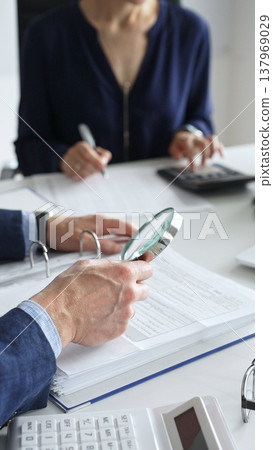 Business colleagues are working together, analyzing financial documents with a magnifying glass, calculating and taking notes in the office. Audit and taxes in business 137969029