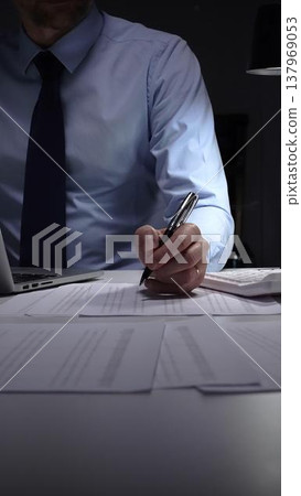 Businessman working meticulously with financial documents, using a pen and calculator to manage accounting tasks and reporting. Audit and taxes in business Businessman working meticulously with financial documents, using a pen and calculator to manage accounting tasks and reporting. Audit and taxes in business 137969053