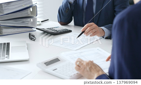 Male accountant analyzing financial documents, calculating numbers together at shared workspace with laptop and office supplies surrounding them. Audit and taxes concept 137969094