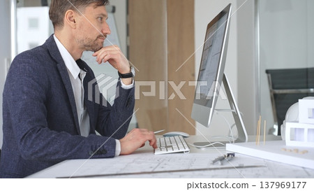 Man architect designing building plans on computer, thinking and concentrating on construction project blueprints at office desk. Business and architecture concept Man architect designing building plans on computer, thinking and concentrating on construction project blueprints at office desk. Business and architecture concept 137969177