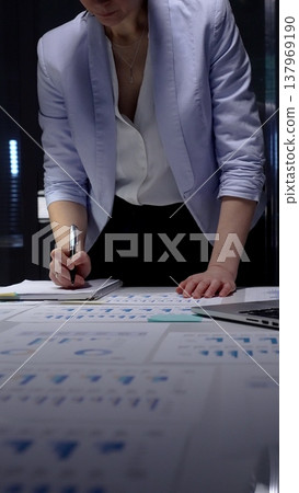 Businesswoman studying financial charts and reports on a desk late at night, analyzing data and strategy for investment, growth and corporate decision making Businesswoman studying financial charts and reports on a desk late at night, analyzing data and strategy for investment, growth and corporate decision making 137969190