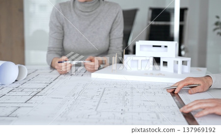 Architects collaborating on architectural design, discussing plans and blueprints with a scale model of a modern house on a desk during a business meeting, close up Architects collaborating on architectural design, discussing plans and blueprints with a scale model of a modern house on a desk during a business meeting, close up 137969192