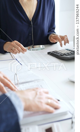 Female financial analysts examining documents, using magnifying glass and calculator while studying corporate data at workplace. Audit and taxes Female financial analysts examining documents, using magnifying glass and calculator while studying corporate data at workplace. Audit and taxes 137969244