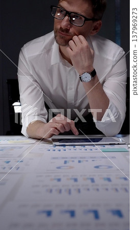 Businessman leans over a desk strewn with charts and reports, working on a tablet in a dim office late at night, reflecting overtime, focus, and relentless dedication 137969273