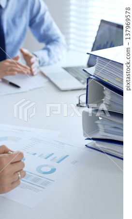 Two professional auditors reviewing financial documents, working collaboratively at white desk with blue folder stack, highlighting teamwork. Audit and taxes in business Two professional auditors reviewing financial documents, working collaboratively at white desk with blue folder stack, highlighting teamwork. Audit and taxes in business 137969578