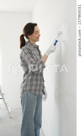 Middle aged happy woman wearing checked shirt, protective gloves, is rolling white paint onto wall, updating living space with fresh color and personal home renovation effort Middle aged happy woman wearing checked shirt, protective gloves, is rolling white paint onto wall, updating living space with fresh color and personal home renovation effort 137969581