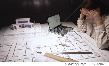 Female architect facing burnout after long late nights, rubbing tired eyes while holding glasses amid blueprints, laptop and a modern building model on her cluttered desk Female architect facing burnout after long late nights, rubbing tired eyes while holding glasses amid blueprints, laptop and a modern building model on her cluttered desk 137969826