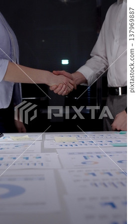 Business partners shaking hands over desk with financial documents, concluding deal, showing success in collaboration, partnership, and agreement during a late meeting at office Business partners shaking hands over desk with financial documents, concluding deal, showing success in collaboration, partnership, and agreement during a late meeting at office 137969887