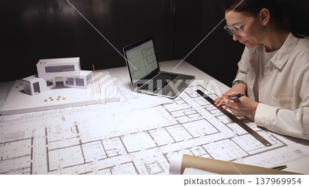 Architect drawing architectural floor plans on a large blueprint paper, working on a complex residential project with a laptop displaying cad designs and a physical house model on the desk Architect drawing architectural floor plans on a large blueprint paper, working on a complex residential project with a laptop displaying cad designs and a physical house model on the desk 137969954