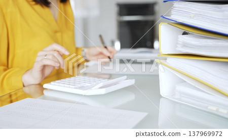 Woman in yellow blouse calculating finances at office desk, using calculator and writing, surrounded by documents and binders. Audit and taxes in business 137969972