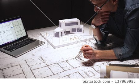 Architect drawing precise measurements and evaluating blueprints while also working on a physical architectural model and reviewing computer aided design plans on a laptop Architect drawing precise measurements and evaluating blueprints while also working on a physical architectural model and reviewing computer aided design plans on a laptop 137969983
