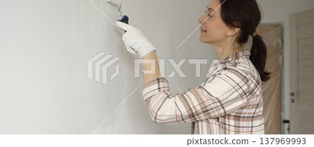 Woman wearing protective gloves painting white wall with small paint roller, updating interior and giving room clean, modern look during home renovation project Woman wearing protective gloves painting white wall with small paint roller, updating interior and giving room clean, modern look during home renovation project 137969993