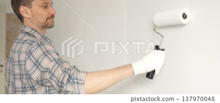 Man professional painter wearing checkered shirt, is rolling white paint on apartment wall with paint roller, transforming interior space with steady strokes during comprehensive home renovation Man professional painter wearing checkered shirt, is rolling white paint on apartment wall with paint roller, transforming interior space with steady strokes during comprehensive home renovation 137970048