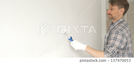 Man professional painter wearing checkered shirt, is rolling white paint on apartment wall, transforming interior space with steady strokes during comprehensive home renovation 137970053