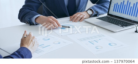 Business colleagues or partners are discussing financial data on a laptop and printed documents while analyzing charts and market trends at office table. Businesspeople and finance concept Business colleagues or partners are discussing financial data on a laptop and printed documents while analyzing charts and market trends at office table. Businesspeople and finance concept 137970064