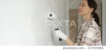 Painter woman wearing gloves is skillfully applying paint to a white wall with a roller in an apartment undergoing renovation, transforming the space with her creative touch Painter woman wearing gloves is skillfully applying paint to a white wall with a roller in an apartment undergoing renovation, transforming the space with her creative touch 137970068