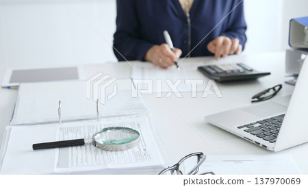 Professional analyzing financial data, writing notes near laptop, magnifying glass, documents, binders on workplace desk. Audit and taxes concept Professional analyzing financial data, writing notes near laptop, magnifying glass, documents, binders on workplace desk. Audit and taxes concept 137970069