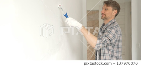 Man professional painter wearing checkered shirt, is rolling white paint on apartment wall, transforming interior space with steady strokes during comprehensive home renovation 137970070