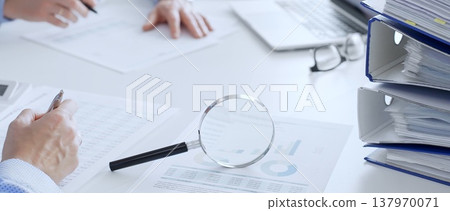 Business people analyzing financial data on charts and documents with laptop and folders on white desk in office, close up of magnifying glass. Audit and taxes theme in business 137970071