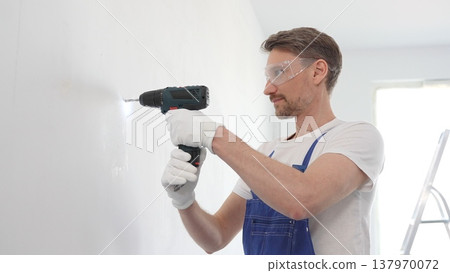 Man professional construction worker in a blue jumpsuit, safety goggles, and work gloves is screwing a screw into the wall using a cordless screwdriver, home renovation 137970072