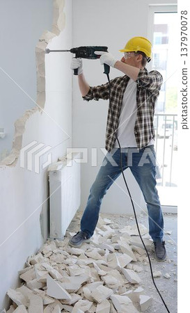 Construction worker demolishing concrete wall with rotary hammer, wearing safety gear, generating rubble and dust during extensive home renovation process 137970078
