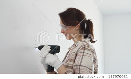 Woman wearing protective gear drilling hole in white wall during home improvement project, focused on precise home renovation work Woman wearing protective gear drilling hole in white wall during home improvement project, focused on precise home renovation work 137970081