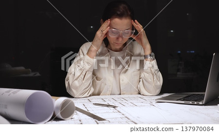 Stressed architect having a headache while working late at night, surrounded by blueprints, laptop, and architectural models, struggling with project deadlines 137970084
