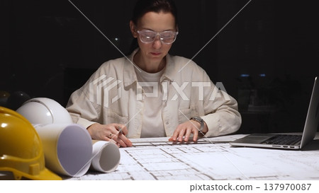 Female architect working late on detailed building blueprints using ruler and pencil, modern engineer designing construction plans at desk with laptop and hardhat 137970087