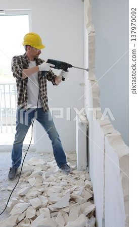 Man construction worker demolishing concrete wall with rotary hammer, wearing safety gear and yellow hardhat, generating rubble and dust during extensive home renovation process 137970092
