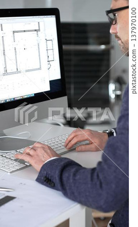 Architect designing building plans on computer, typing at desk with architectural model and blueprints. Business and architecture concept 137970100