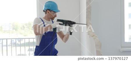 Male construction worker demolishing concrete wall with rotary hammer, wearing work overalls and blue hardhat is generating dust and debris during home renovation work, portrait horizontal view 137970127