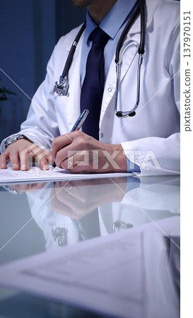 Medical professional wearing a white lab coat. Stethoscope. And tie. Sitting at a glass desk. Calculating paperwork. A laptop visible on the side Medical professional wearing a white lab coat. Stethoscope. And tie. Sitting at a glass desk. Calculating paperwork. A laptop visible on the side 137970151