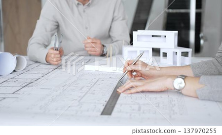 Architectural team analyzing building plans and a white scale model, discussing design concepts and construction details during a professional meeting in an office environment, close up 137970255