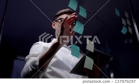 Man in eyeglasses organizing thoughts and ideas for business optimization on a clear glass board. Creating a strategic roadmap for innovation and project planning Man in eyeglasses organizing thoughts and ideas for business optimization on a clear glass board. Creating a strategic roadmap for innovation and project planning 137970297
