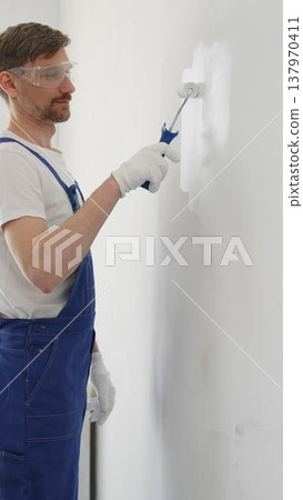 Man professional painter wearing protective white gloves and blue work overalls, applying paint with roller on pristine interior wall during home renovation project, vertical view Man professional painter wearing protective white gloves and blue work overalls, applying paint with roller on pristine interior wall during home renovation project, vertical view 137970411