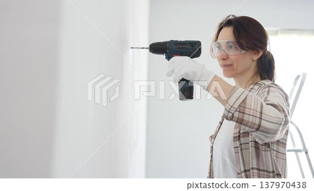 Middle aged happy woman wearing protective gear and gloves, is drilling hole in white wall during home improvement project, focused on precise home renovation work 137970438
