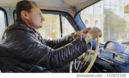 Male driver riding on blue vintage auto at city road on sunny day. Serious man driving retro vehicle at urban street. Confident guy moving in his old auto on route. Slow motion Male driver riding on blue vintage auto at city road on sunny day. Serious man driving retro vehicle at urban street. Confident guy moving in his old auto on route. Slow motion 137972109