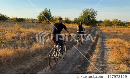 Young male friends ride mountain ski bikes along trail on sunny summer day. Beautiful golden sunlight at background. Concept of active lifestyle, adventure spirit and carefree weekend outdoor Young male friends ride mountain ski bikes along trail on sunny summer day. Beautiful golden sunlight at background. Concept of active lifestyle, adventure spirit and carefree weekend outdoor 137972119